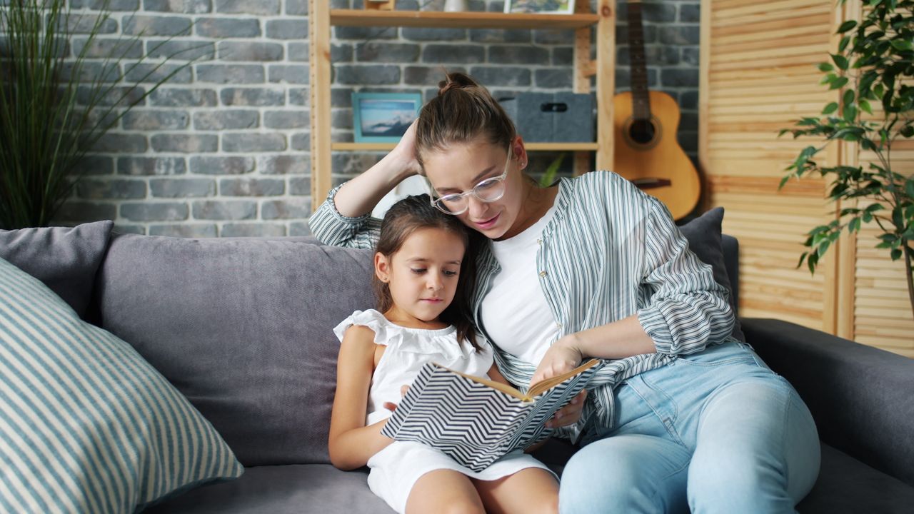 A mother and daughter enjoying quality time reading a book on the sofa indoors.